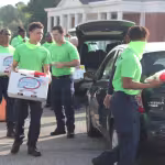 Volunteers in bright green shirts unload boxes from a car during a community event. The scene is busy, with a collaborative and energetic mood.