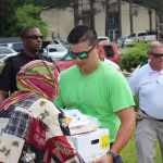 A man in a green shirt and sunglasses hands a box of supplies to a person in a colorful headscarf at an outdoor community event, surrounded by volunteers.