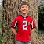 A smiling child in a red football jersey with the number 2 stands against a large tree trunk in a wooded area, conveying a cheerful and playful mood.