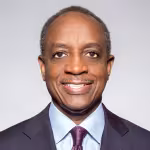 A man in a dark suit with a light blue shirt and patterned tie smiles warmly against a plain light gray background. The image conveys a professional tone.