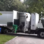 A garbage truck lifts a green bin to empty its contents. A worker in a yellow vest operates the truck. A blue recycling bin is visible nearby, surrounded by leafy trees.
