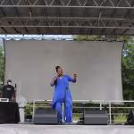 A woman in a vibrant blue outfit energetically sings on an outdoor stage. Two men stand to the left near sound equipment. Trees are visible in the background.