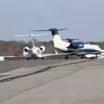 Private jets line up on a runway, with trees in the background under a clear blue sky, conveying a sense of luxury and travel readiness.