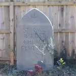 A stone monument with a cross and the words "St. Paul Baptist Church Cemetery" stands in front of a wooden fence, surrounded by overgrown plants.