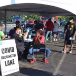 Outdoor COVID vaccination site under a tent, where a healthcare worker in uniform administers a vaccine to a seated woman. Sign in foreground.
