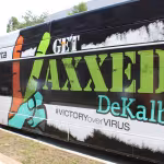 Side view of a bus with colorful lettering "Get Vaxxed DeKalb" and "#VictoryOverVirus", parked on a sunny street, promoting vaccination.