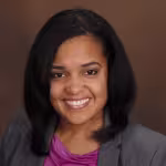 A woman with shoulder-length black hair smiles warmly. She is wearing a gray blazer over a purple shirt against a simple brown background.