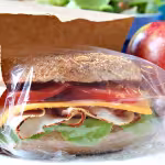 A whole grain sandwich with lettuce, sliced turkey, cheddar, and tomato in plastic wrap peeks from a brown bag, next to a shiny red apple on a table.