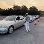 A line of cars waits in a parking lot, organized with orange traffic cones. A man in casual attire speaks to a driver. The sky is overcast.