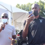 Two men wearing masks and sunglasses stand outdoors. One speaks into a microphone, while the other holds up a smartphone. A large tent is in the background.