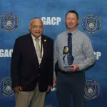 Two men stand smiling in front of a blue GACP backdrop. One man wears a suit, the other holds an award, wearing a checked shirt and tie.