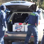 Two people in blue uniforms with "RECRUIT" on the back load supplies into an SUV. A large box labeled "Cheryl's Best" is seen. Scene is outdoors.