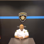 Person in a white uniform sits at a desk, flanked by US and Georgia flags, under a DeKalb County Marshal badge. The room feels official and formal.