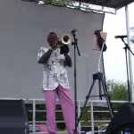 A musician in colorful attire plays the trumpet on an outdoor stage, with a mic and a tablet on stands nearby, conveying a vibrant, live performance.