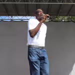 Tall man in a white shirt and jeans speaking into a microphone on an outdoor stage, under a clear blue sky, conveying a confident and engaging presence.