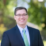 A man in a suit and green tie smiles warmly outdoors. The background is blurred with green trees, suggesting a sunny, pleasant day.
