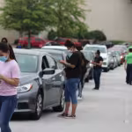 People wearing masks stand beside a line of parked cars, engaging with drivers. The scene depicts a drive-thru event on an overcast day.