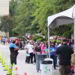 People wearing masks line up outside under trees next to a building, supervised by a police officer. Cones guide them, hinting at an organized event.
