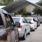 A line of cars waits under a large tent at an outdoor event. A dog looks out from a truck window, adding a curious and eager vibe to the scene.