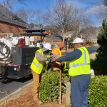 Three workers in high-visibility vests and helmets dig near shrubs, next to a utility truck on a residential street. Sunny day with trees and houses.