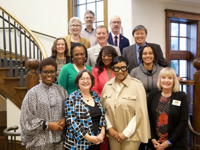 A diverse group of twelve people pose on a staircase, all smiling warmly. The setting is formal, with wooden railings and large windows.