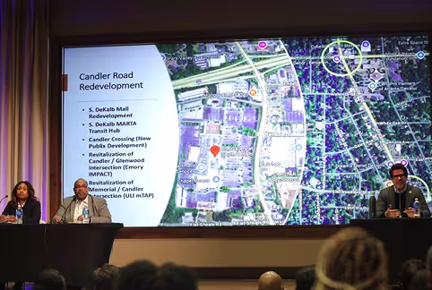 Three speakers sit at tables on stage in front of an audience at the DeKalb County Q3 Town Hall. Behind them, a large screen displays a map and a list titled Candler Road Redevelopment, aiming to engage residents in key project areas.