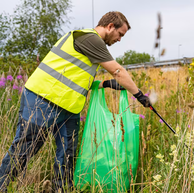 A local man participates in the Adopt-a-Mile program, wearing a Hi-Viz vest as he picks up trash with a grabber and puts it in a green bag on the side of the road.