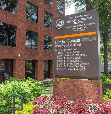 A Large brown sign with an orange stripe in front of a red brick building and green, red, and pink shrubbery that reads "DeKalb County Government Manuel J. Maloof Center Administrative Building & Auditorium". It lists the name of the CEO and its board commissioners.