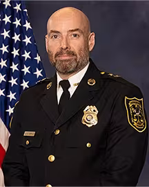 Man in traditional Police dress uniform with a gold badge on his chest with an American flag in the background.