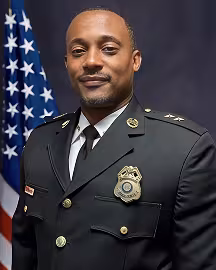 Man wearing a black traditional dress uniform with a gold badge, gold buttons, and a gold pin on either collar in front of the American flag and a black background.