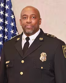 Man wearing a black traditional dress uniform with a gold badge, gold buttons, and a gold pin on either collar in front of the American flag and a light tan background.