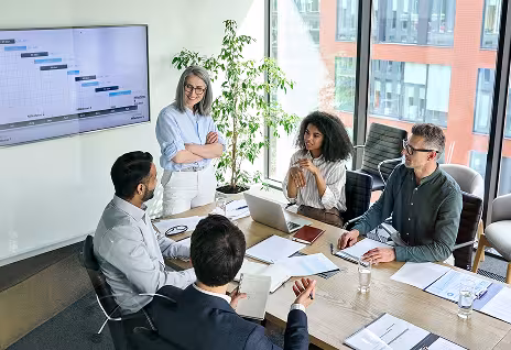 Five people meeting around a desk, four are sitting, listening to a presentation on a television screen behind them.