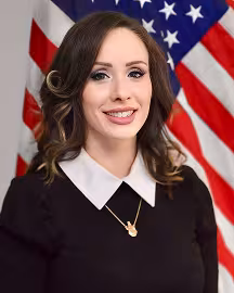 Public Education Specialist Brittany Godfrey, with long brown hair and a black dress with a white collar smiles in front of a U.S. flag. The tone is formal and professional.