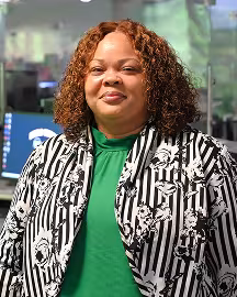 Woman with brownish red hair wearing a green shirt and a black and white vertical striped jacket with a flower pattern.