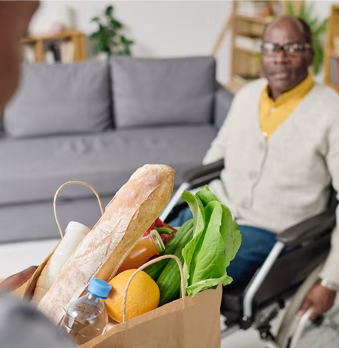 Man in a yellow shirt and a beige cardigan sitting in a manual wheelchair with a brown paper bag of groceries on a table.