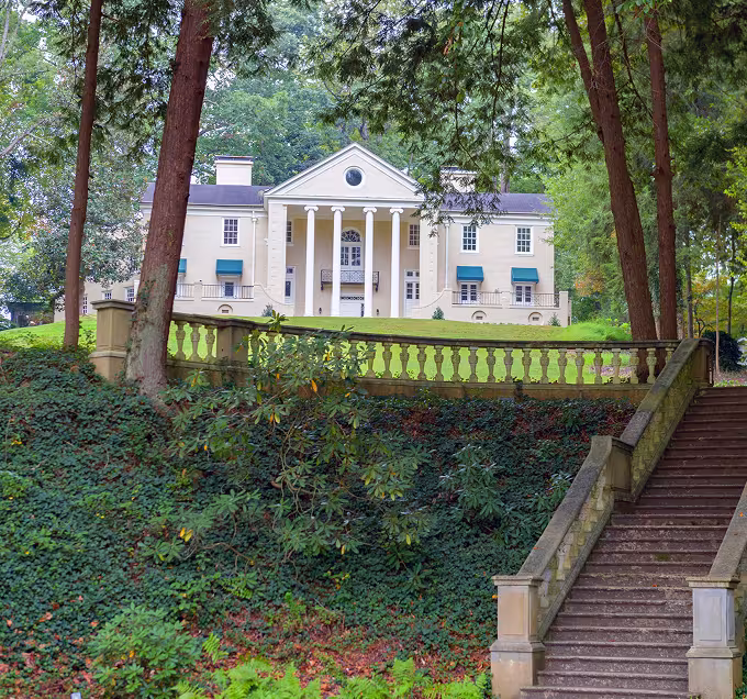 Elegant manor house with large columns, surrounded by greenery. Stone staircase leads up to the house, exuding a serene and historic ambiance.