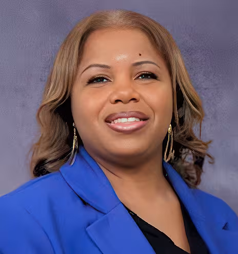 A woman with light brown hair, hooped earrings, and a blue blazer with a black shirt, smiling in front of a grey background.