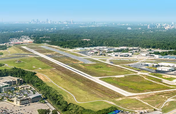Wide landscape photo of a long runway surrounded by green grass with an airport in the background.