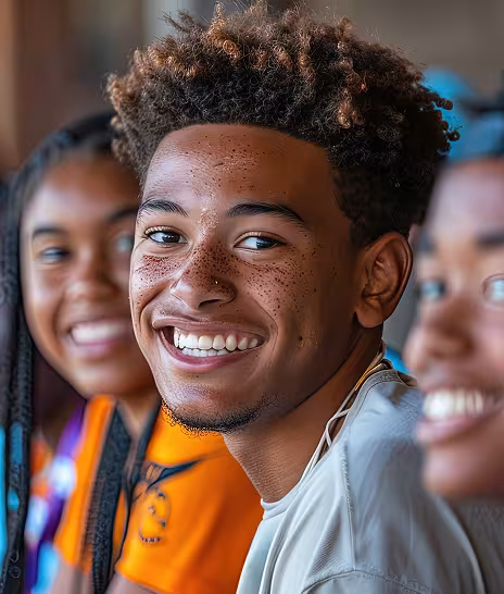 Young man with tall brown hair and a grey shirt with a young woman behind him in an orange shirt. Both are smiling.