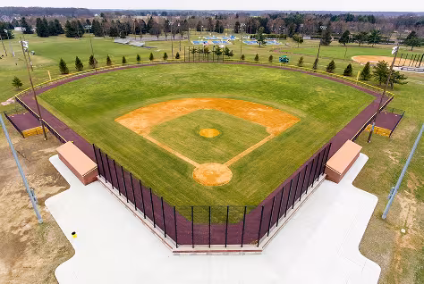 Aerial view of an empty DeKalb County baseball field with neatly mowed grass, brown warning track, and surrounding fencing, plus additional sports fields and trees in the background.