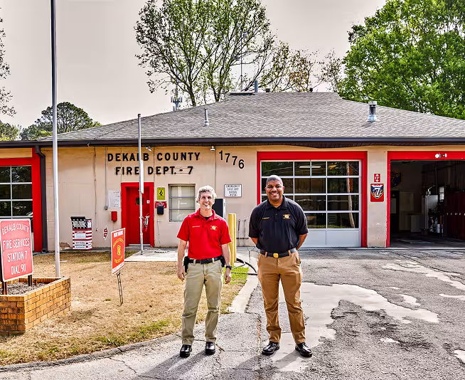 Two men stand in front of DeKalb County Fire Department Station 7, with the SPLOST I-funded fire station building and garage doors visible in the background.