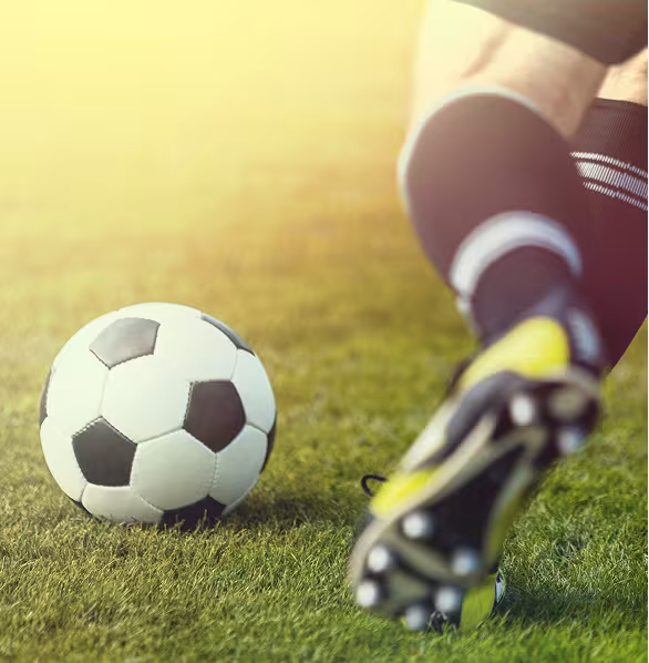A soccer player wearing black knee-high socks kicks a soccer ball on a lush green field. The sun casts a warm, golden glow over the scene.