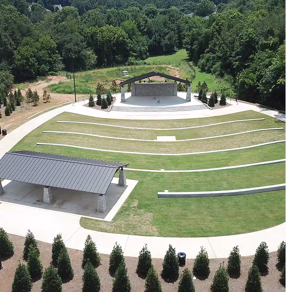 An aerial view of an outdoor amphitheater surrounded by lush trees. It features a covered stage, tiered seating on grass, and a nearby pavilion. Peaceful and inviting.