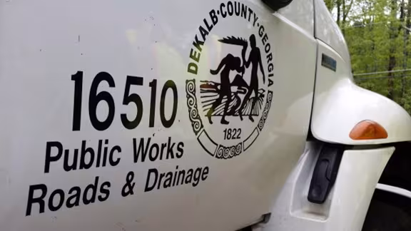 White truck with "DeKalb County, Georgia" seal and text "16510 Public Works Roads & Drainage." Forest background suggests a roadside location.