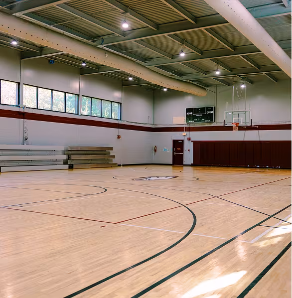 Spacious indoor basketball court with polished wooden floors and red and black lines. Bleachers on the left, hoop and scoreboard on the right. Quiet atmosphere.