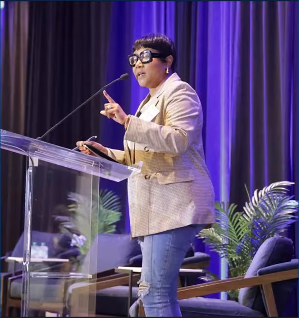 A person speaks passionately at a podium with a microphone, gesturing with one hand. They wear a blazer and jeans. The background is lined with potted plants and deep blue curtains.