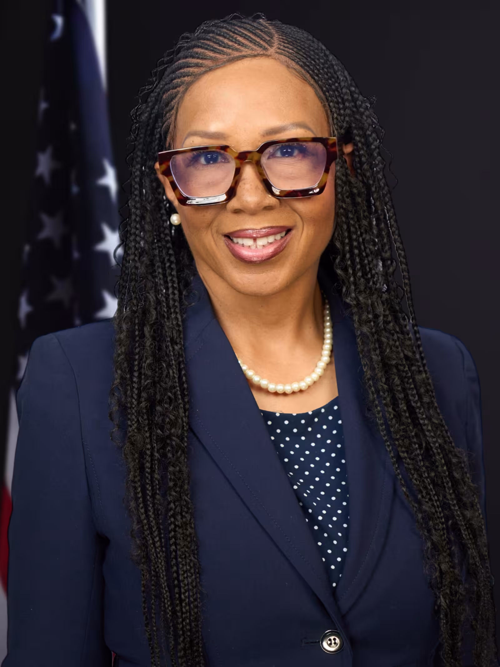 A woman with long braided hair and tortoiseshell glasses is smiling. She wears a navy blazer, polka dot blouse, pearl necklace, and pearl earrings. An American flag is partially visible in the background.