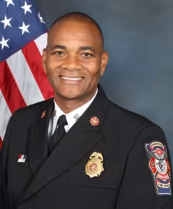 Smiling man in a firefighter dress uniform with badges and patches, standing in front of an American flag. The background is a plain blue-gray.