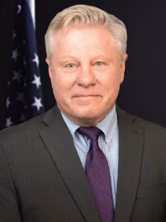 Man in a suit with a purple tie stands against a dark background. A blurred U.S. flag is visible behind him. He appears serious and professional.
