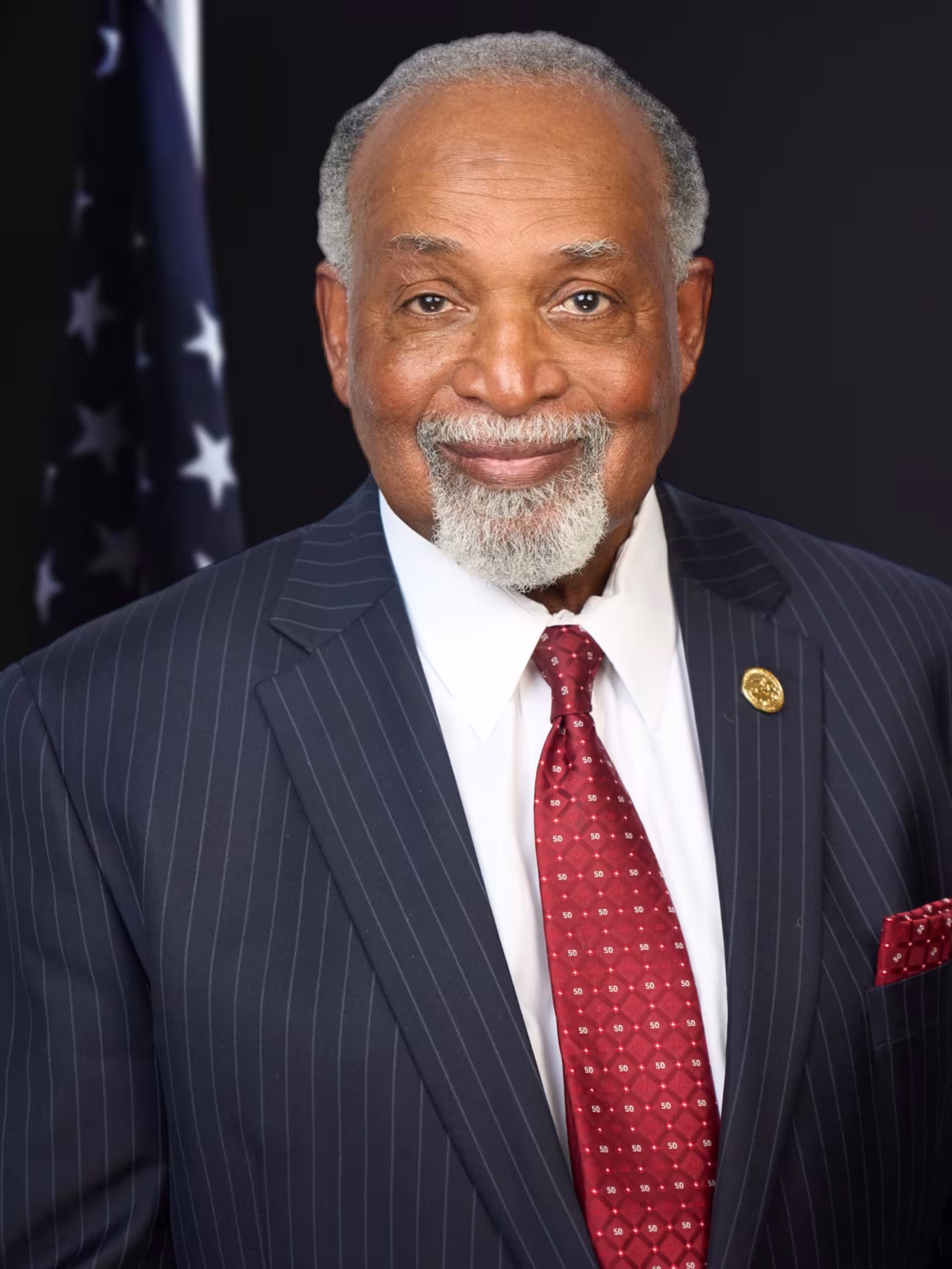 An elderly man smiles warmly, wearing a dark pinstripe suit, white shirt, and red patterned tie, against a dark background with a subtle American flag.